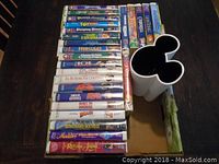 25 Disney and Pixar VHS movies in original cases laid out on a dark wood table with Mickey Mouse shaped vase