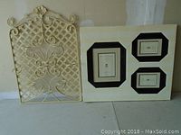 Metal candle holder and picture frames decor lot placed on floor against wall. Candle holder with lattice pattern and scrollwork, picture frame has three black-bordered photo openings in white frame.