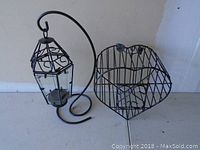 Two black metal decor pieces side by side: a hanging lantern holder with a curved top and a heart-shaped wall decor with vertical bars and decorative swirls