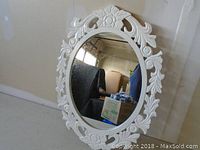 White oval mirror with ornate white frame leaning against a wall, showing detailed floral and scroll design on frame.