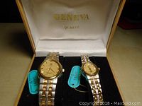 Two Geneva quartz watches, a larger men's style and a smaller women's style, with gold and silver linked metal bands inside a white lined presentation box.