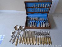 Full set showing silver plated and gold plated cutlery pieces arranged outside and inside the wooden box with blue lining.