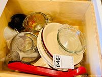 Drawer with various kitchen items including measuring cups, pie dishes, strainer, funnel, tongs