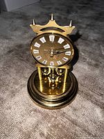 Front view showing brass clock, crown finial, textured dial with Roman numerals and four-ball pendulum