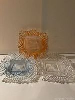 Front view of three square pressed glass dishes in clear, amber-peach and light blue