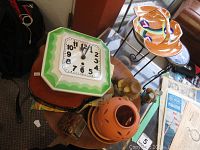 Photo of 8 day wall clock on a small table with orange ceramic lantern, small wooden sculptures, and other decor items nearby.