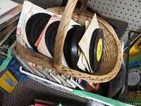 Basket of multiple 45 RPM vinyl records mostly in paper sleeves, displayed side by side, showing various colors and label designs on the records