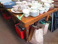 Overall view of oak table used as a surface for stacked teacups and plates, showing the table top and leg design with surface scratches visible.