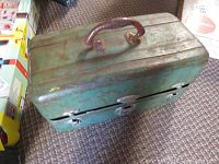 Closed green metal fishing tackle box on carpet, showing handle and latch closures.