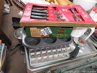 Photo showing various bakeware items stacked including muffin tin, rectangular aluminum baking sheets, a metal tray with multiple square molds, and a pastry press box on top with knives visible.