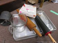 Aluminum baking sheets stacked with wooden rolling pins, boxed muffin tin, and Fire-King measuring cup visible on top.
