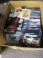 Box filled with approximately 60 DVDs and PC DVD-ROM games. Visible titles include action and comedy genres, loose and stacked inside a cardboard box.