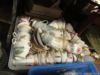 Photo of a large box filled with dozens of mismatched teacups and saucers, mostly white porcelain with floral and gold designs.