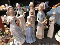 Photo showing 6 lady porcelain figurines in a group, some holding flowers, birds, or baskets, on a display with other collectibles in background.