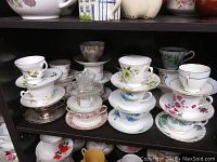 21 assorted tea cups and saucers arranged in four stacks on black shelving showing various floral patterns and gold trim.
