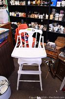 Photo of vintage painted wood high chair, white color, set in a cluttered room with various ceramics and collectibles in the background.