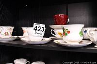 Seven vintage teacups and saucers displayed on a black shelf, various floral patterns and one red cup visible.