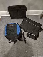 Three bags laid out: blue/black Lowepro backpack, black MEC laptop sleeve, black messenger bag