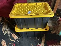 Two black plastic totes stacked, showing yellow lids and ribbed sides