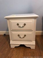 Front view of white wooden nightstand side table with two drawers and decorative handles on a wood floor against a gray wall.