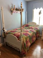 Twin size white wooden trundle bed in a bedroom with four tall posts, covered with a multicolored quilt and striped bed skirt. Bed is against blue walls and near a window.