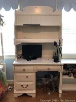 Photo shows white wooden desk with hutch, four drawers including one with broken handle. Hutch has two shelves. Desk is against windows with items on top including a monitor and lamp, which are not included.