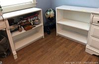 Two white laminate bookcases positioned on a wooden floor near a window and a white dresser. One bookcase contains items on the shelves, the other is empty.