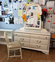 Front view of white wooden dresser with ornate attached mirror and matching upholstered chair next to it, showing all drawers and chair seat.