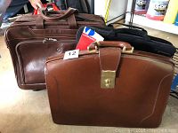 Three briefcases grouped together showing front brown leather briefcase with brass lock, second brown leather case, and black laptop bag visible in the background