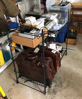 Black wire rack with two shelves containing various items and a brown leather briefcase at the bottom, located in a cluttered garage space.