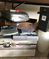 Wide shot of ioSafe hard drive box, Brother label maker, clear plastic tray, spiral notebooks, and Epson scanner on shelf