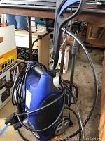 Blue and black pressure washer with power cord coiled, wheels, and handle, shown standing on a concrete floor.