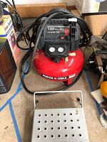 Full view of the air compressor showing red tank, black control top with pressure gauges, and attached air hose.