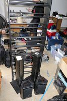 Black metal media rack with horizontal bars, two black pentagonal Tannoy speakers and two black speaker stands positioned in front, all items sitting on concrete floor.