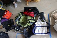 Various soft carry-on luggage bags and a portable folding travel seat photographed on concrete floor with some shoes visible in background.