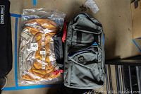 Photo showing one gray Tenba camera bag and one packaged orange Kelty backpack side by side on a floor.