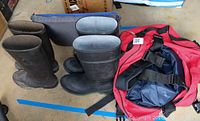 Two pairs of men's outdoor rain boots, one size 7 and one size 9, black color, placed on a ground surface next to a red North Face outdoor duffle bag with black straps.