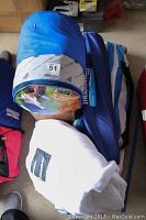 Photo showing 2 folding chairs in blue carry cases, one is rolled and has a label with number 51, placed beside a white rolled sleeping bag