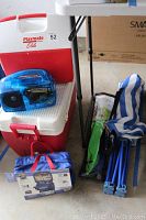 Photo showing Igloo Playmate Elite cooler with red and white plastic, blue see-through radio placed on top, emergency first aid kit and part of folding chair frame and tackle kit bag on the floor.