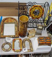 View of various decor items on table including small mirror with wood frame, three handheld mirrors, two table clocks, decorative jewelry box, cylindrical vase, and black wrought iron magazine rack with mounted colorful round plate.