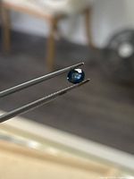Round faceted sapphire held by tweezers showing blue-green hue
