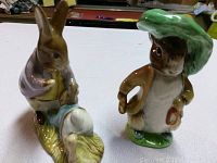 Photo showing two porcelain bunny figurines on a white table: Mr. Benjamin Bunny and Peter Rabbit with a smaller blue-jacketed rabbit