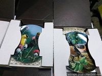 Photo showing two decorative bird plates in original protective cardboard inserts with part of bird illustrations visible.