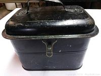 Top and side view of black metal lunchbox showing hinged lid, latch, metal handle, and wear on surface.