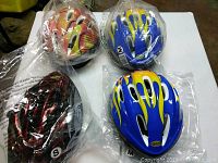 Four bicycle helmets on a table, each in plastic packaging. Three helmets show a blue and yellow flame graphic design and are size medium. One helmet is smaller, black with red flames, size small.