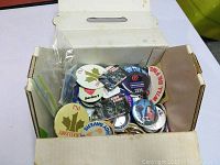 Box filled with assorted collectible buttons and pins, showing various designs and logos.