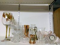 Photo showing the full lot of assorted glassware items on a white shelf including painted wine goblets, crystal glasses, a glass decanter, pink clock, and metallic decorative pieces.