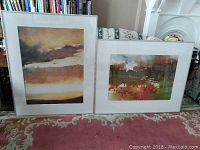 Two large framed abstract prints side-by-side on a floral carpet in front of white fireplace and bookshelf