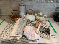 Full lot view: copper kettle, white thermos, two floral teapots, two cookbooks, stack of assorted kitchen linens