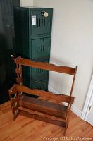 Full view of a tall green metal ammunition locker with four compartments stacked vertically, locked, next to two wooden gun racks placed on the floor.